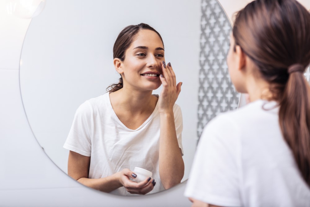 Mulher consciente da importância dos cuidados com a pele olhando-se no espelho e aplicando um dermocosmético no rosto.